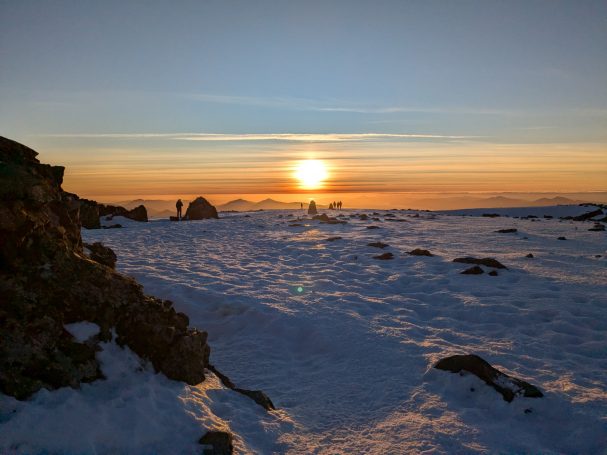 Summit of Ben Nevis Sunset over a snowy landscape with distant silhouettes of mountains and rocks.