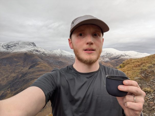 Relaxing before the Summit of An Gearanach Man holding a container, standing on a mountain with snowy peaks in the background.